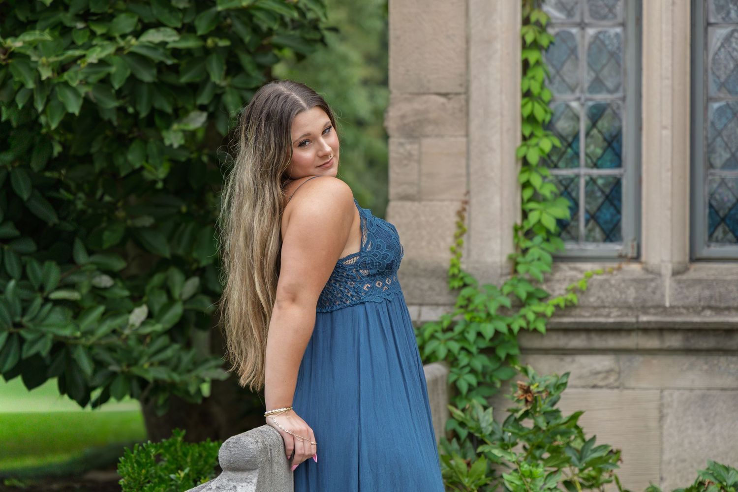 Woman in blue formal gown looking over shoulder near stone building with ivy-covered walls.