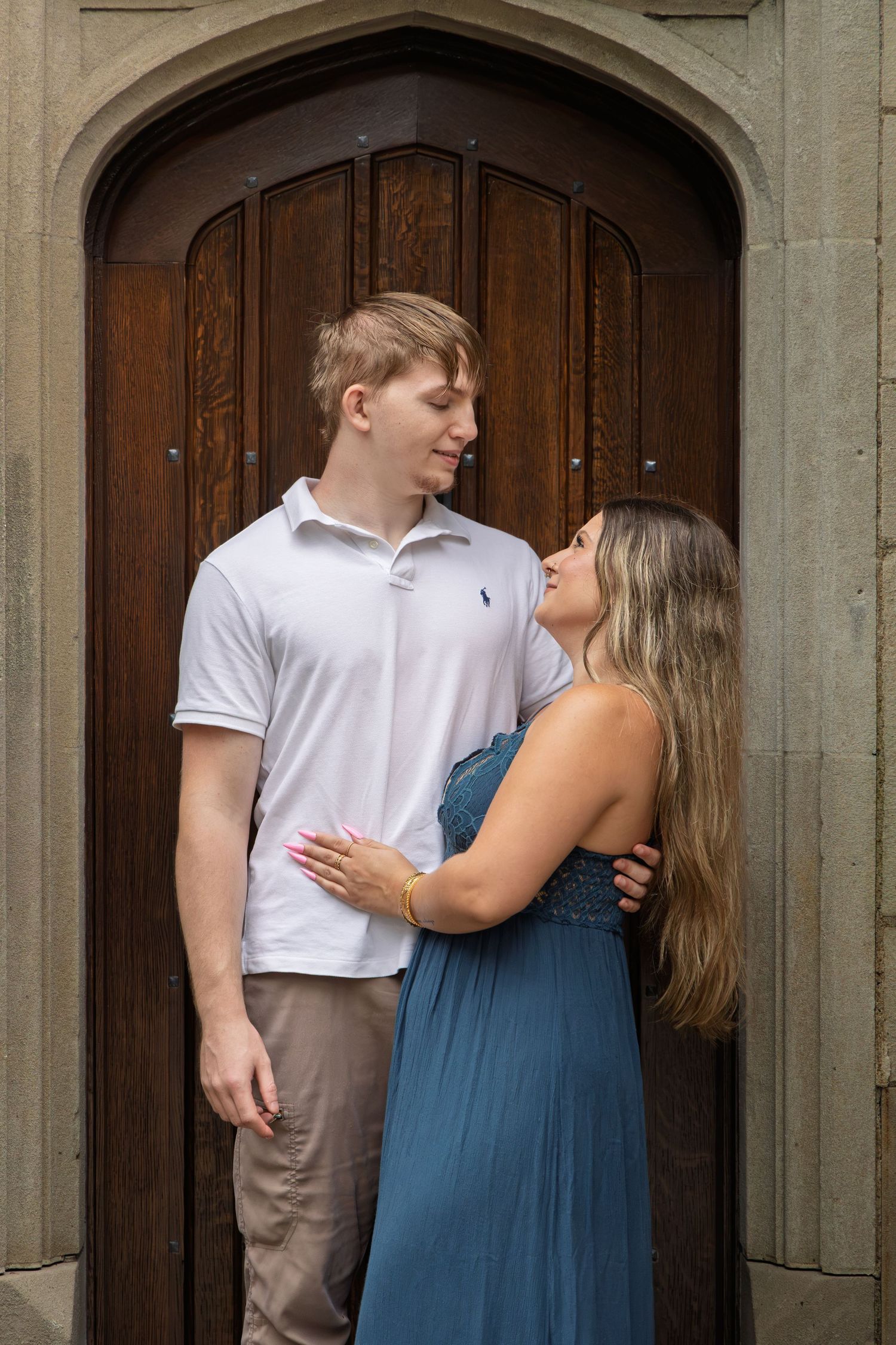 A young couple gazes at each other lovingly in front of a large wooden arched door, she in a blue dress, he in a white polo.