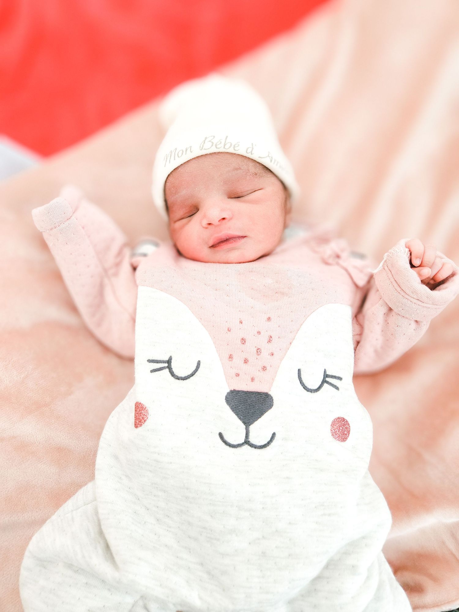 Newborn baby sleeping in a fox-themed swaddle blanket and white knit hat, arms raised up.