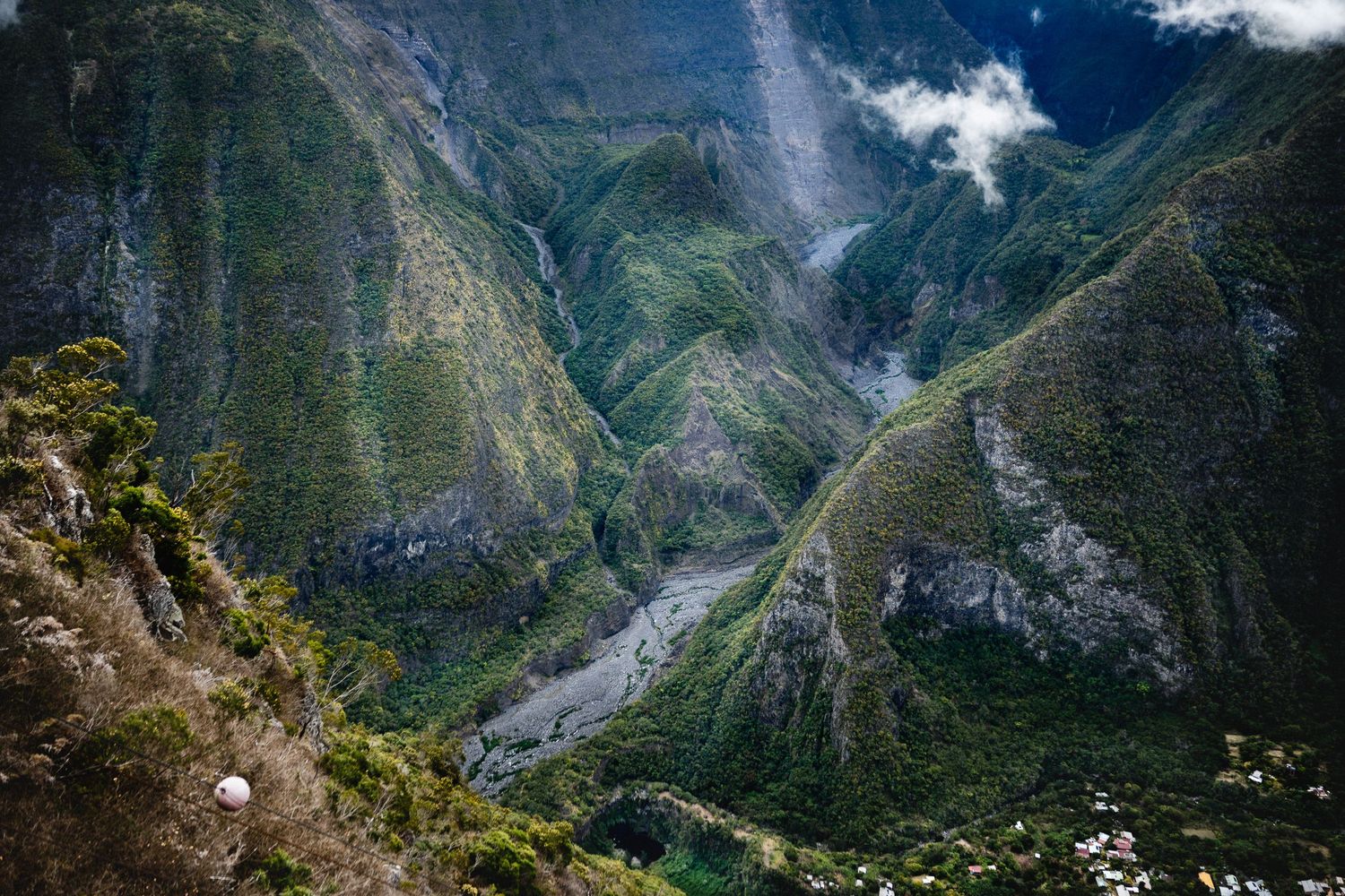 Aerial view of a lush green mountain valley with a winding river and dramatic cliffs under cloudy skies.