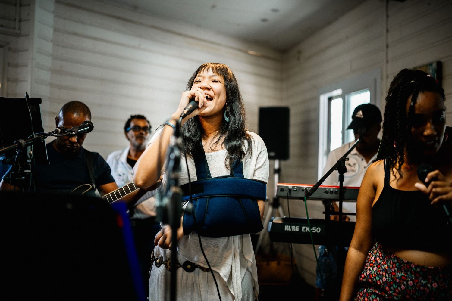 Female singer performing on stage with arm sling while band plays behind her indoors.