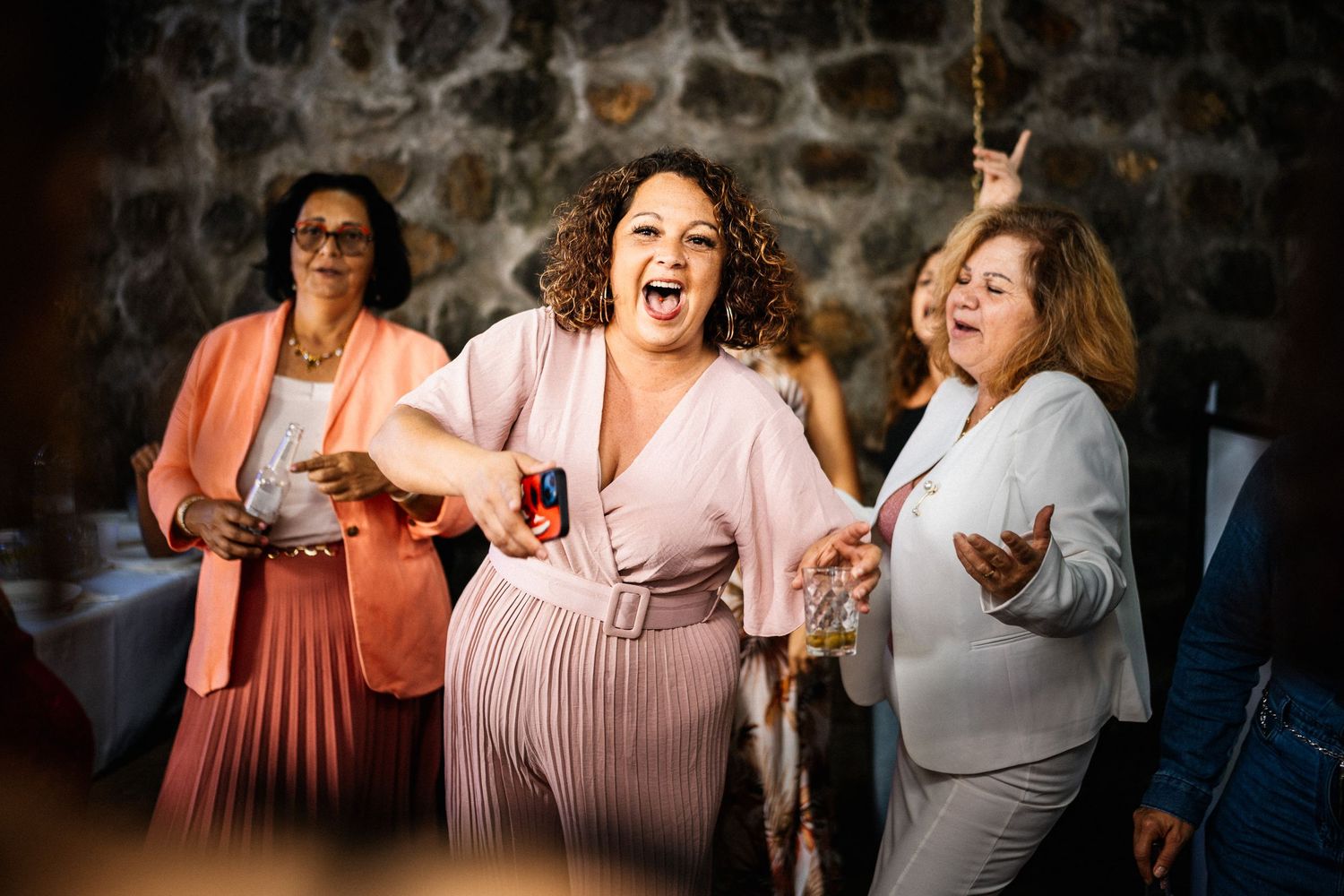 Woman in pink dress laughing joyfully while dancing at a festive party with stone wall backdrop.