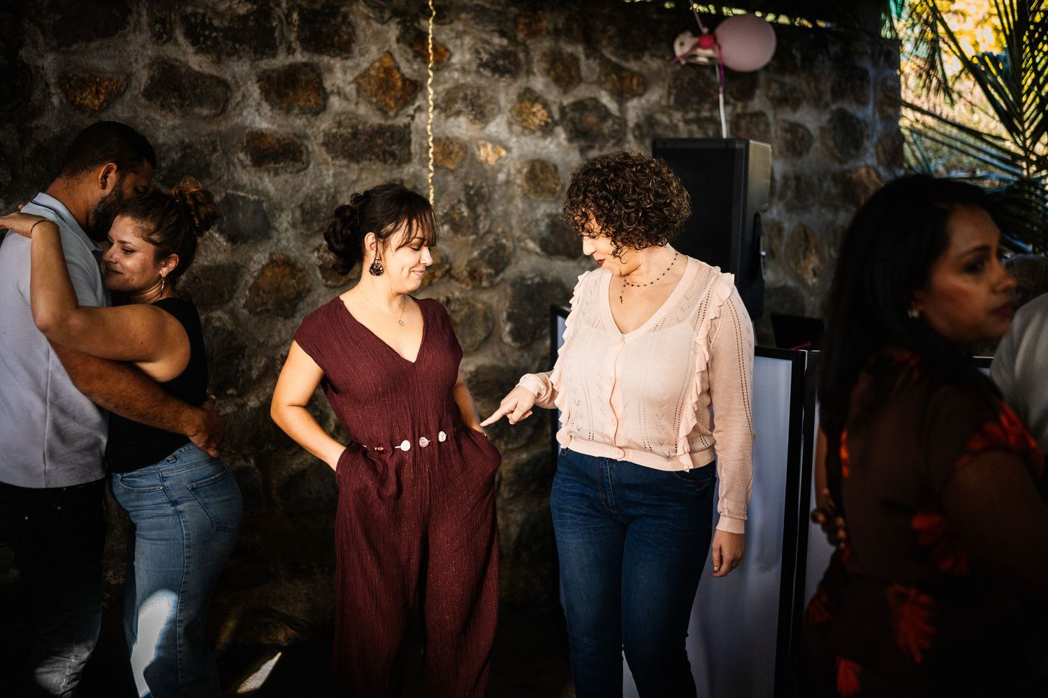 Two women conversing and dancing together at an indoor party with stone wall background.