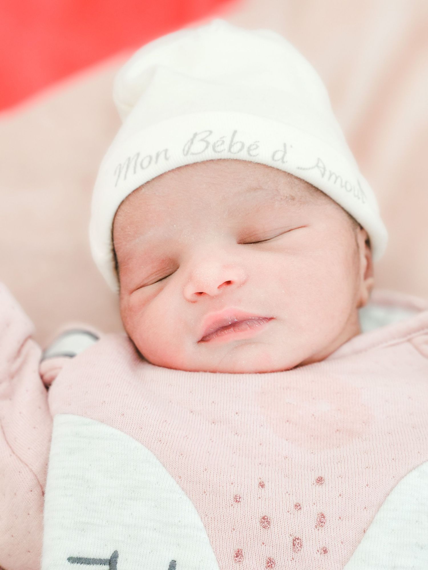 Peaceful newborn baby sleeping in pink outfit wearing white hat reading Mon Bebe d Amour.