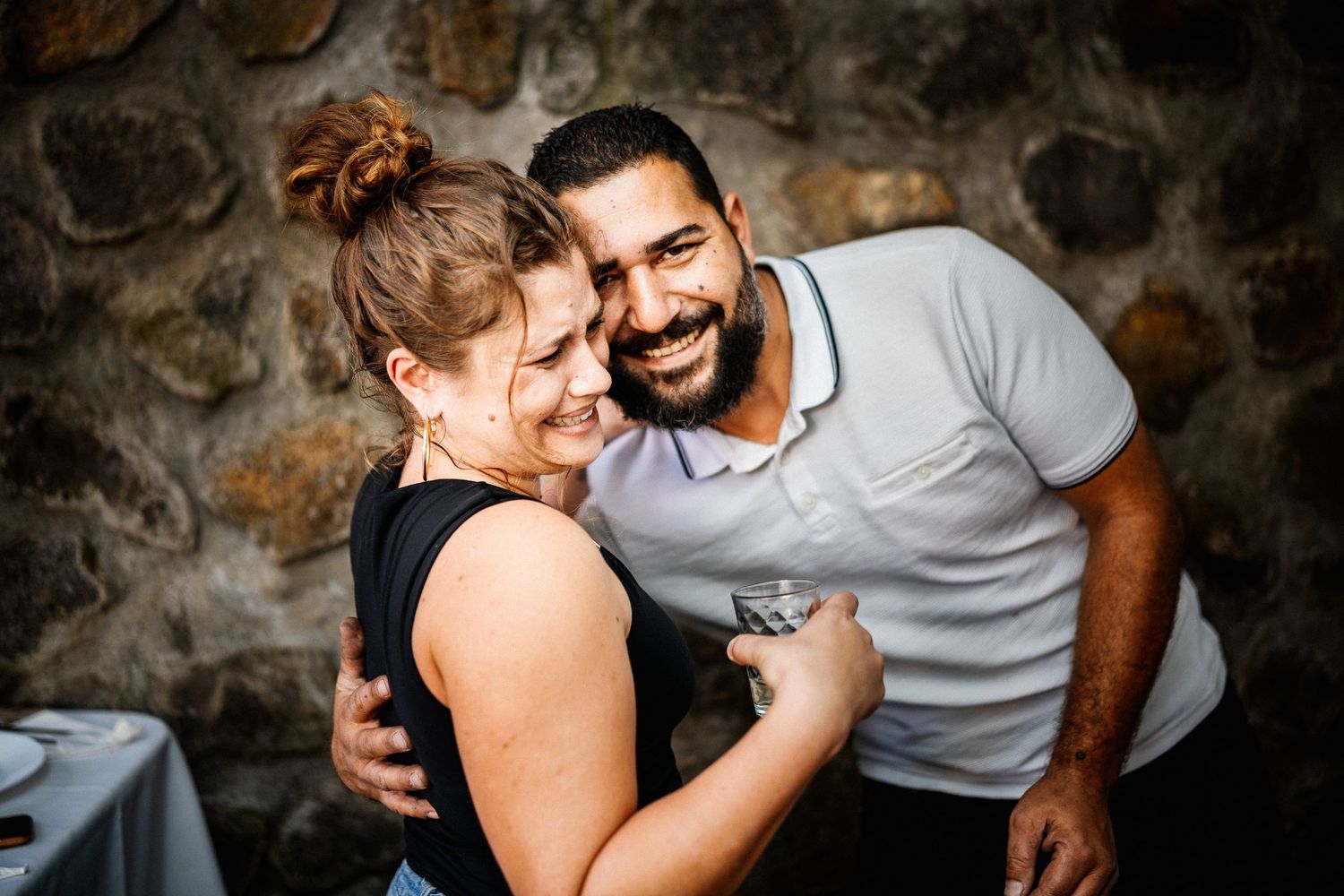 Happy couple embracing and smiling in front of a stone wall at a celebration event.