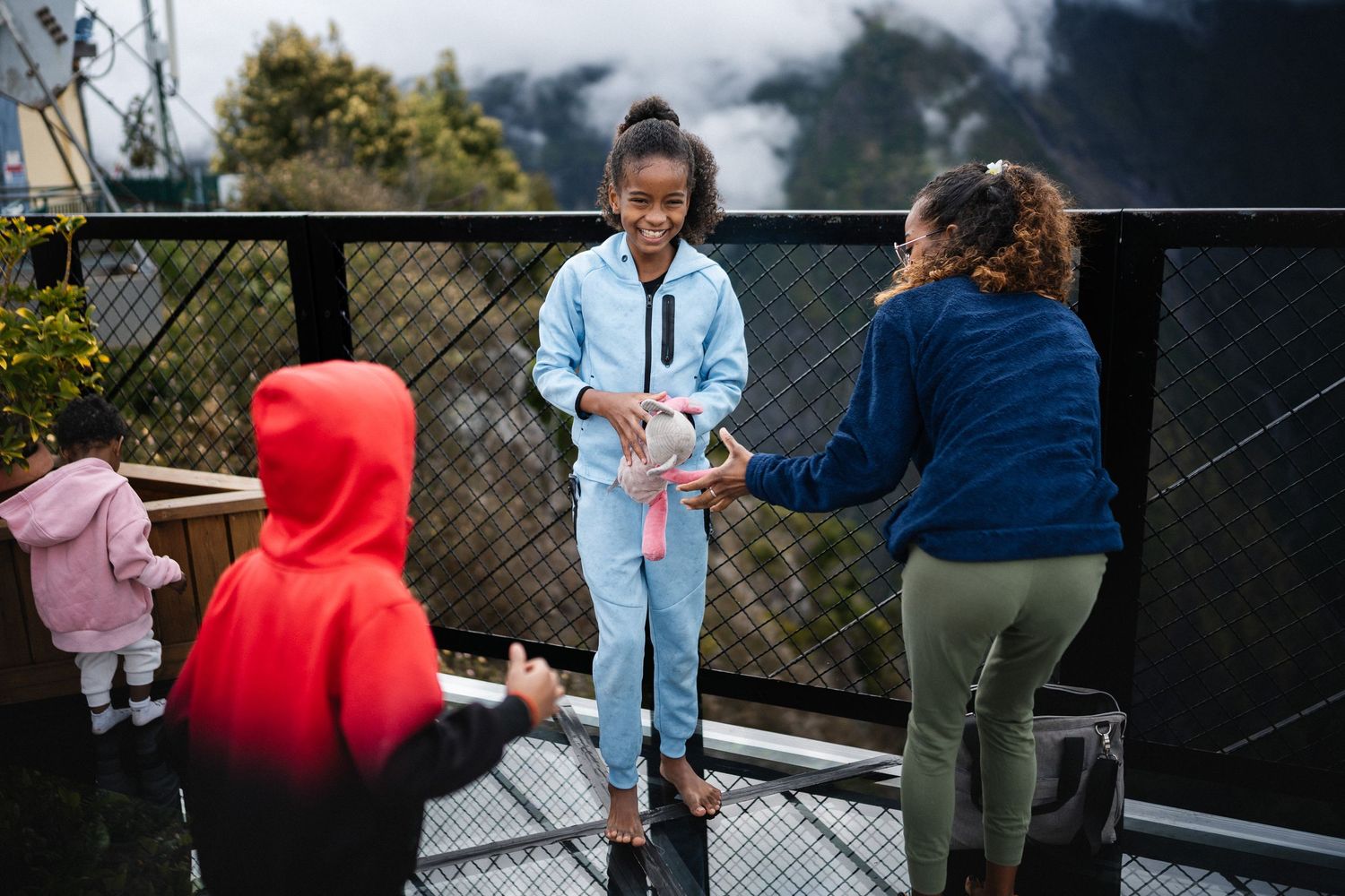 A woman hands a gift to a smiling girl on an outdoor balcony while two other children play nearby in a scenic mountain setting.