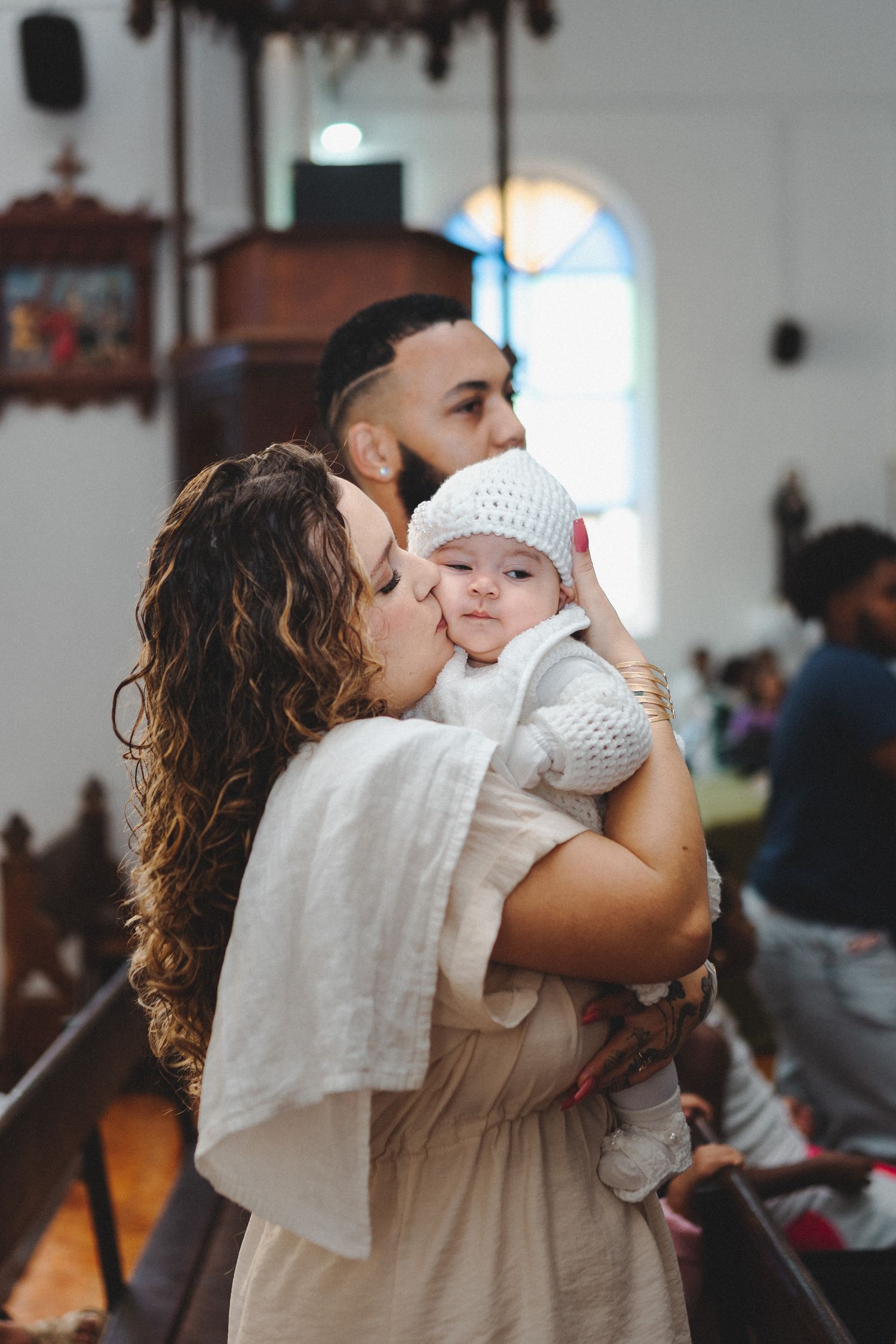 A mother kisses her baby dressed in white at a church baptism ceremony, with the father standing behind them.
