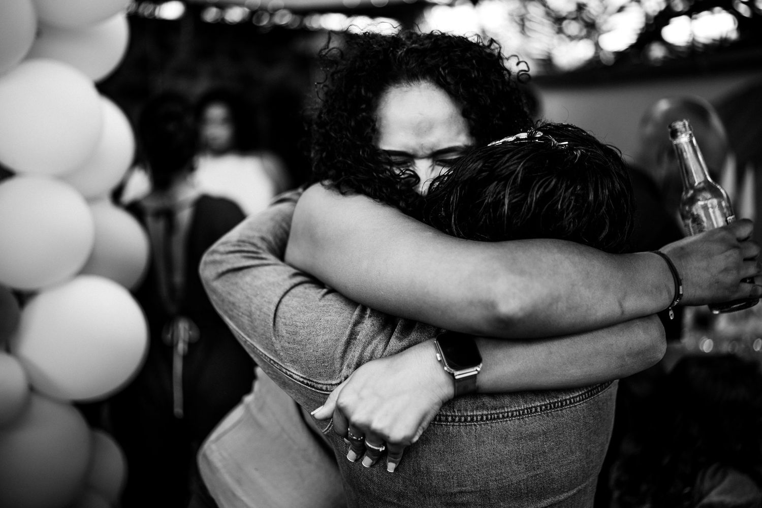 Two people sharing a warm emotional hug at a celebration with balloons in the background.