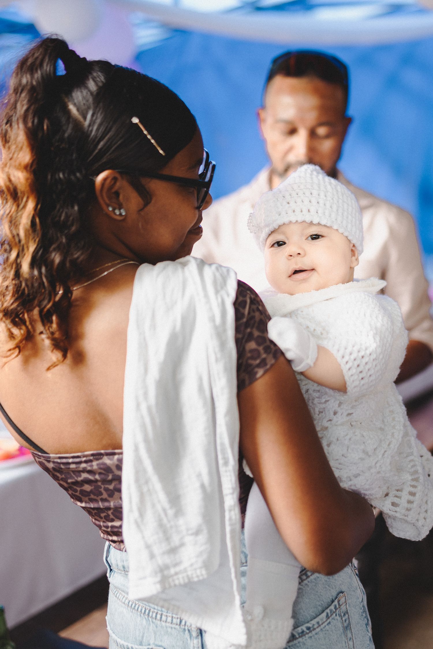 A woman in glasses holds a baby dressed in white crochet outfit, with a man standing in the background.