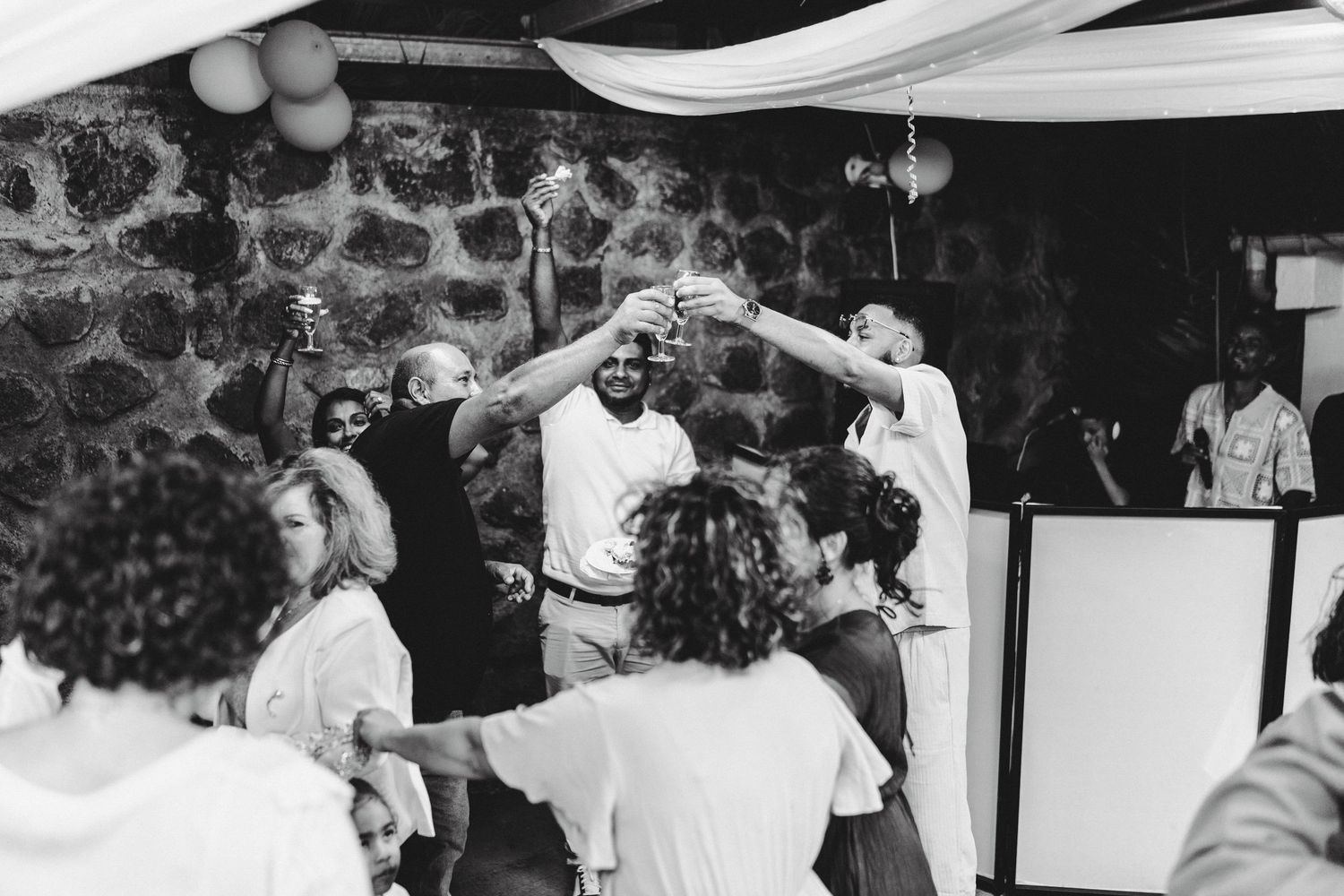 Black and white photo of guests dancing joyfully at a wedding reception in a rustic stone venue.