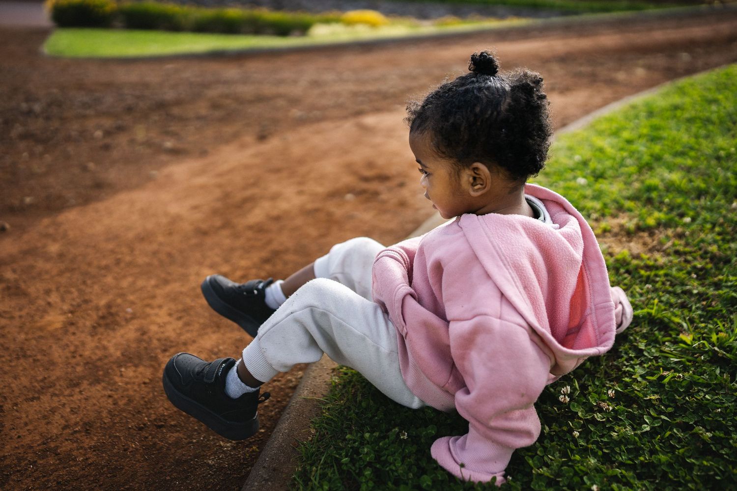 A young Black girl in a pink hoodie and gray pants sits on a grass curb at a playground, looking away thoughtfully.