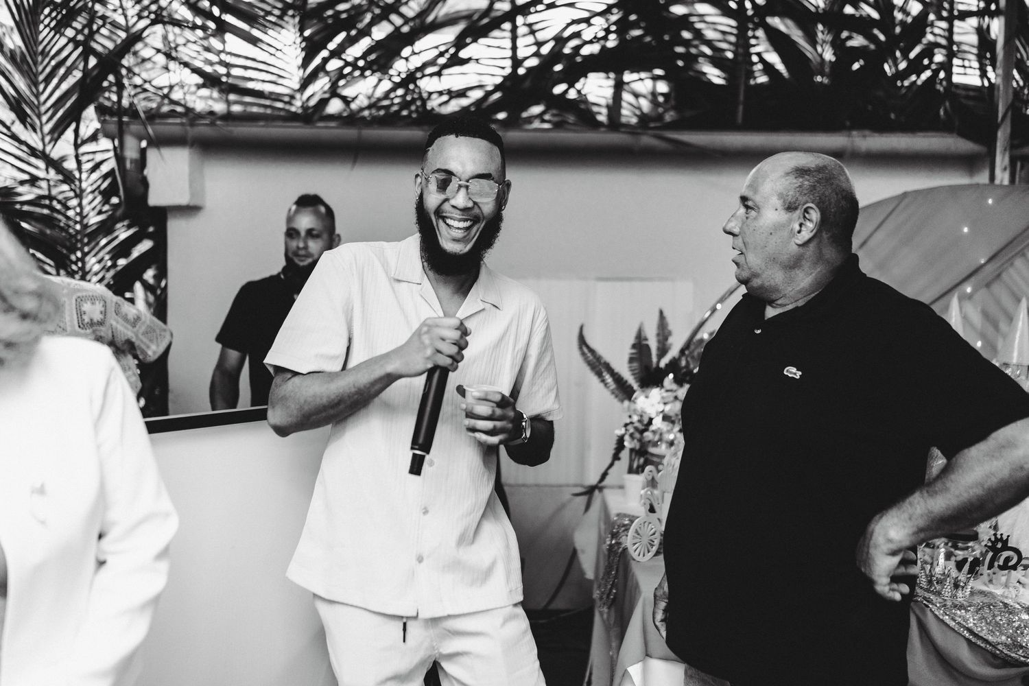 Black and white photo of a laughing man in white outfit holding a mic at an outdoor social gathering with guests.