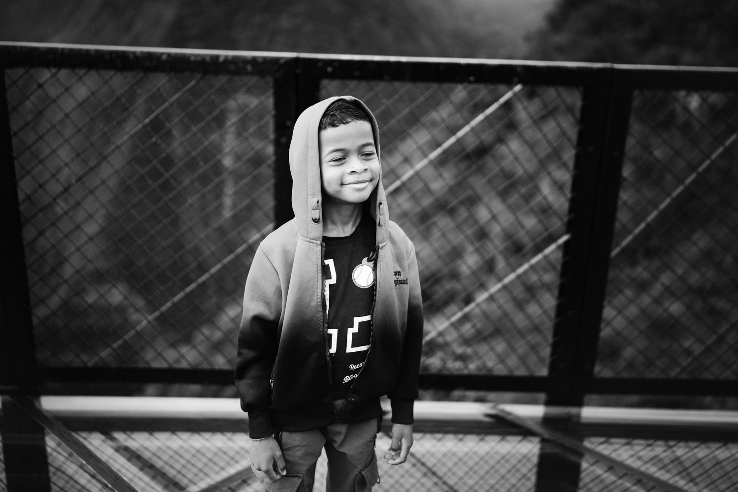 Black and white photo of a smiling boy in a hooded sweatshirt standing on a bridge outdoors.