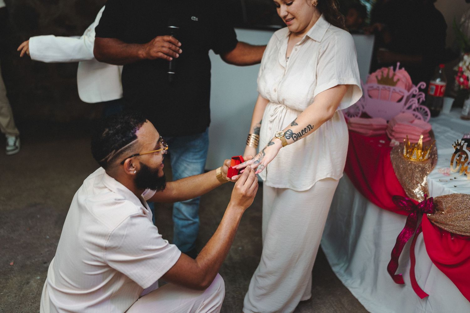 Man kneeling to place a red bracelet on a woman's wrist during an outdoor ceremony with guests nearby.
