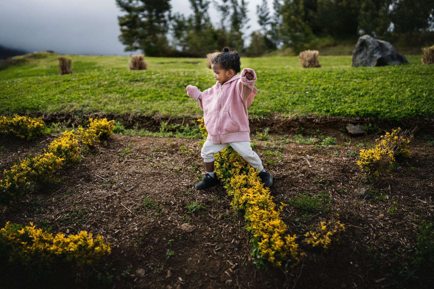 A young girl in a pink hoodie runs through a lush green garden with yellow flowers and hay bales in the background.
