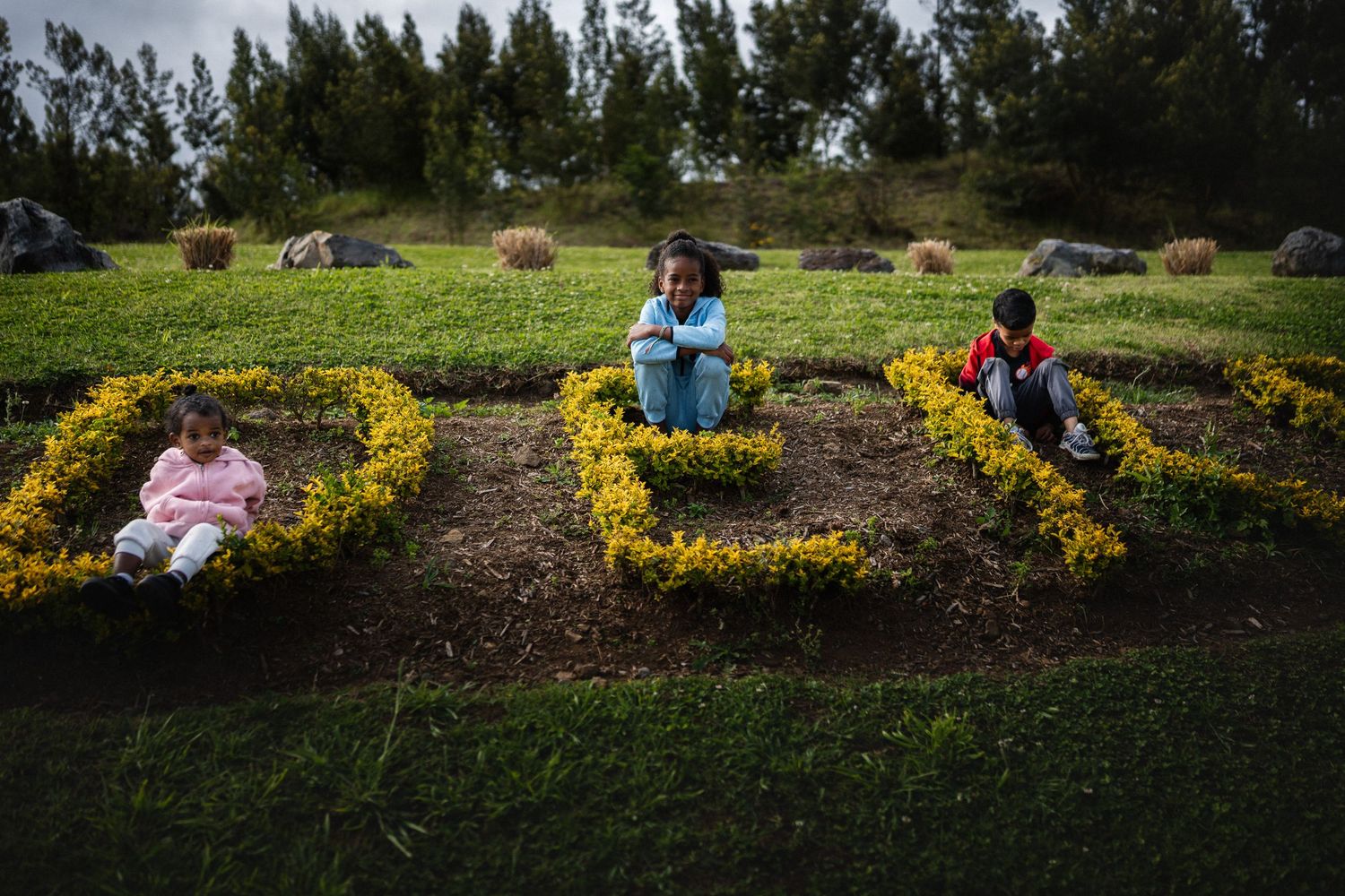 Three children sitting among yellow flower arrangements shaped like letters on a green hillside garden.