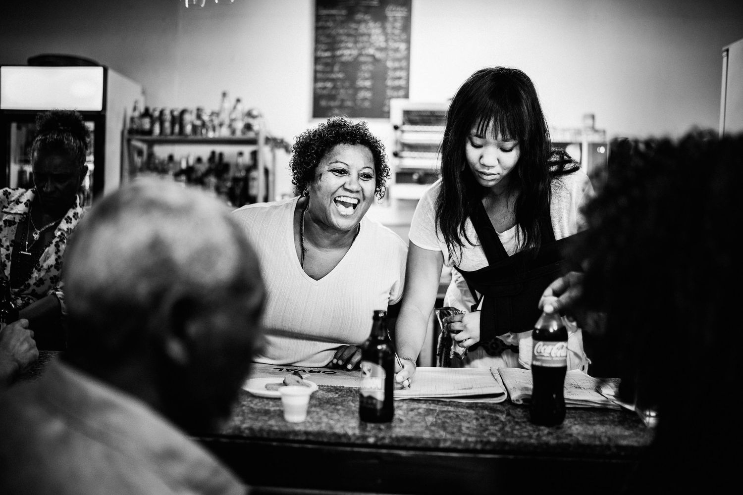 Two women laughing together at a bar counter in black and white candid photo.