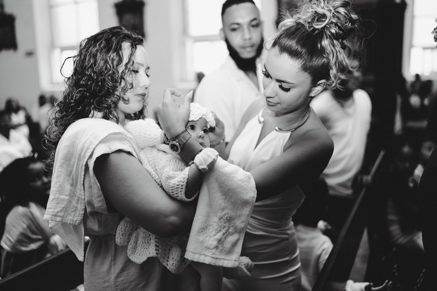 Black and white photo of two women holding a baby at a christening ceremony with guests in the background.