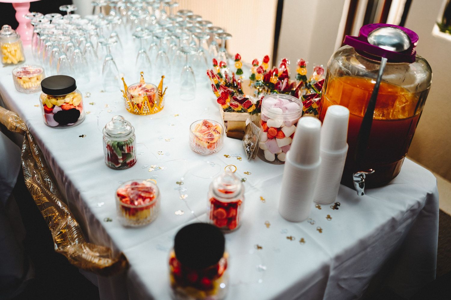 Elegant candy bar table with colorful sweets, desserts, drink dispensers, and white cups at a party event.