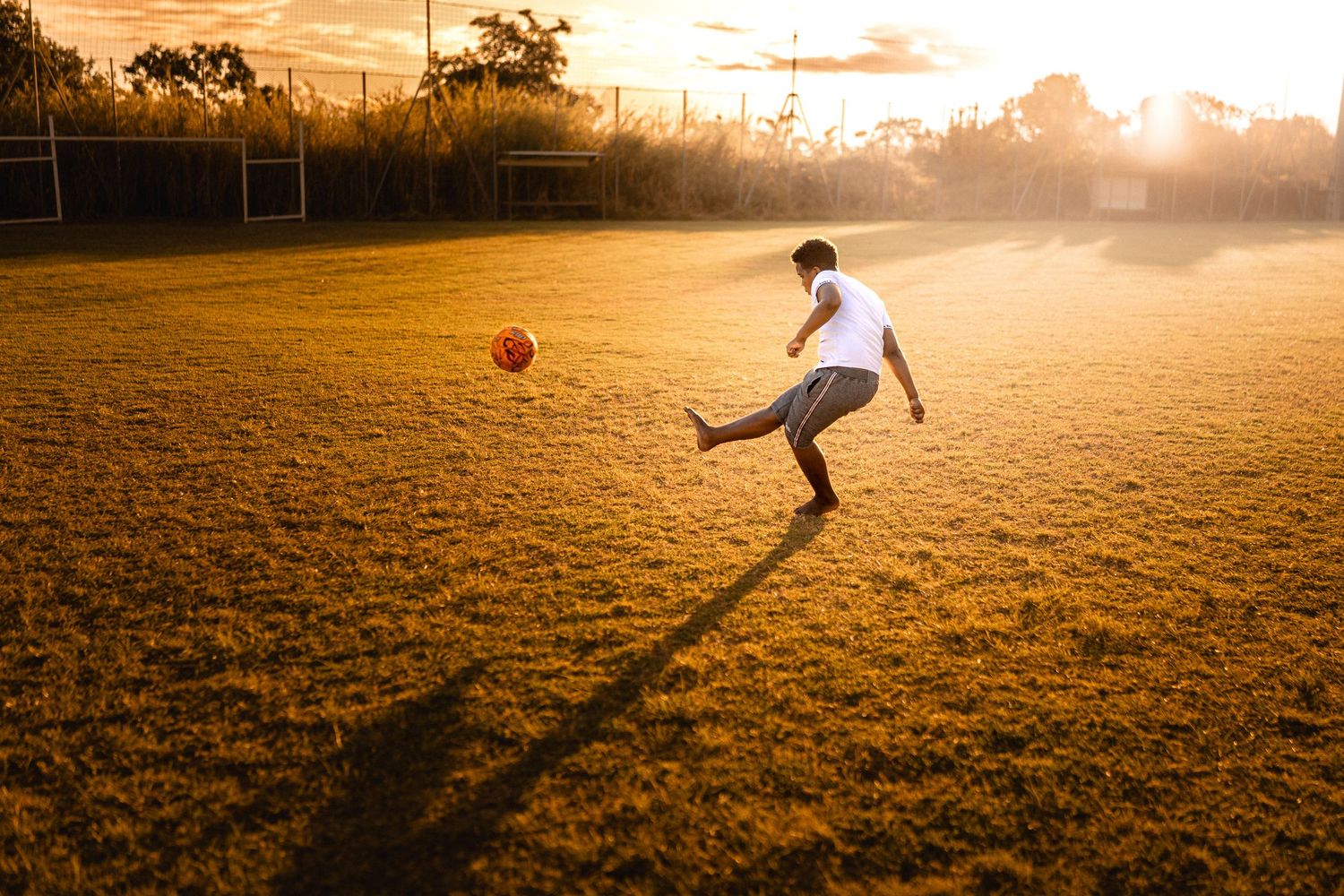 Person kicking a soccer ball on a grass field during a golden sunset, casting long shadows across the pitch.