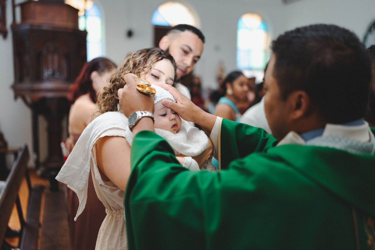 Priest performing a Catholic baptism ceremony on a baby inside a church, with family members gathered around.