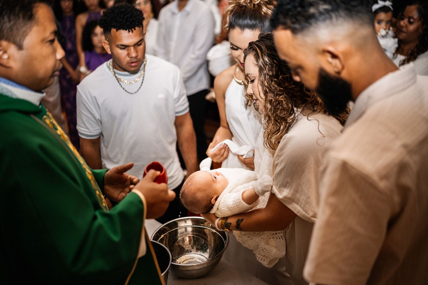 Catholic priest performing a baptism ceremony with family gathered around a baptismal font.