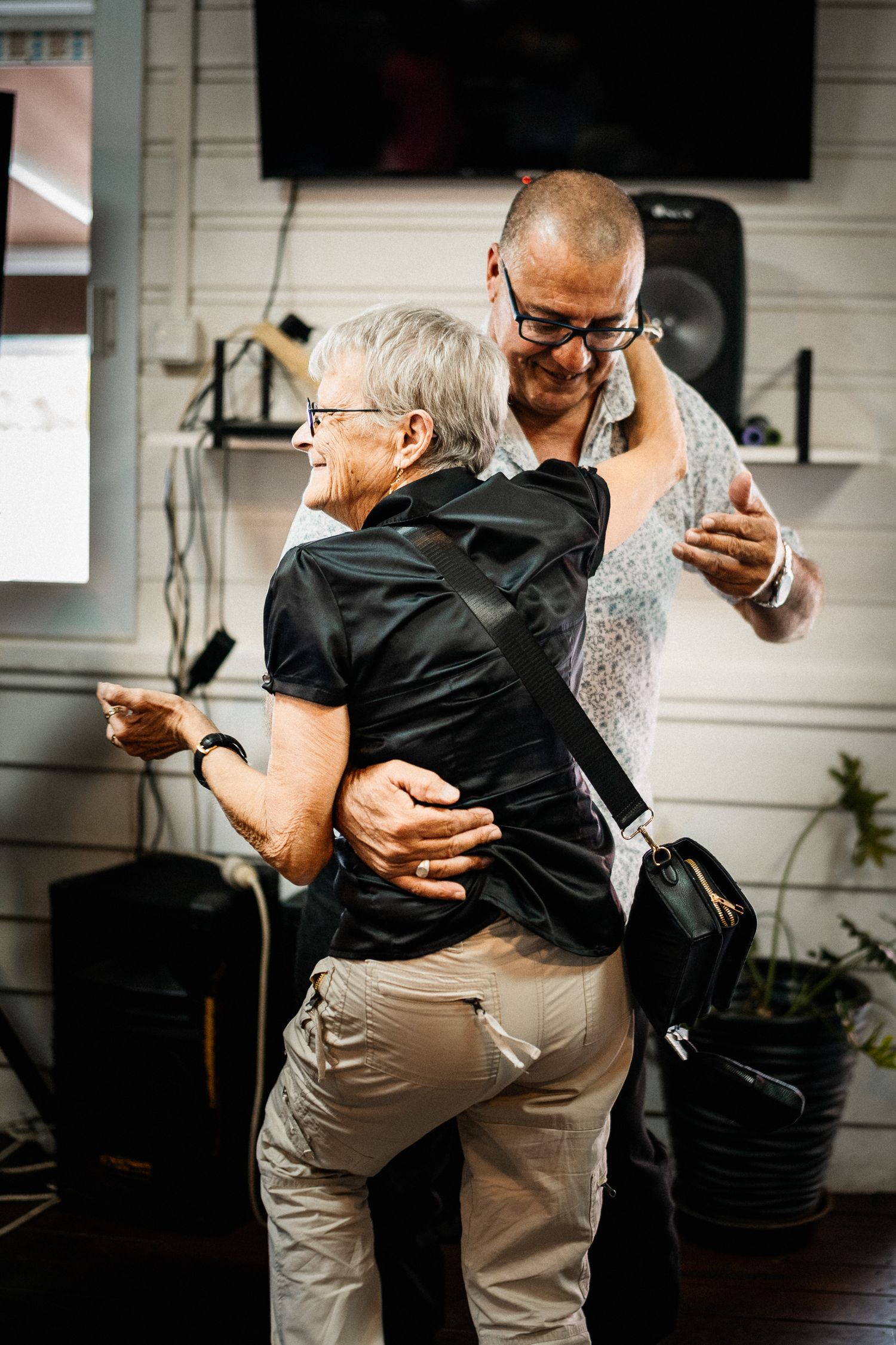 Elderly woman and bald man with glasses sharing a warm dance embrace indoors.