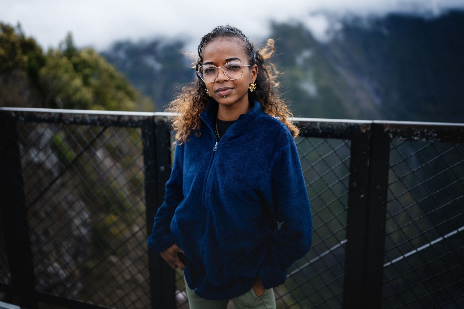 Young woman in blue fleece jacket poses at mountain overlook railing with misty peaks in background.