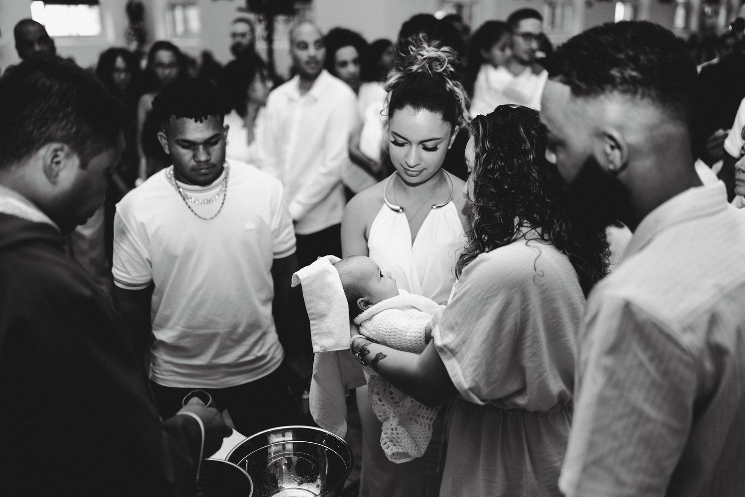 Black and white photo of a baptism ceremony with adults gathered around an infant being held by a smiling woman.