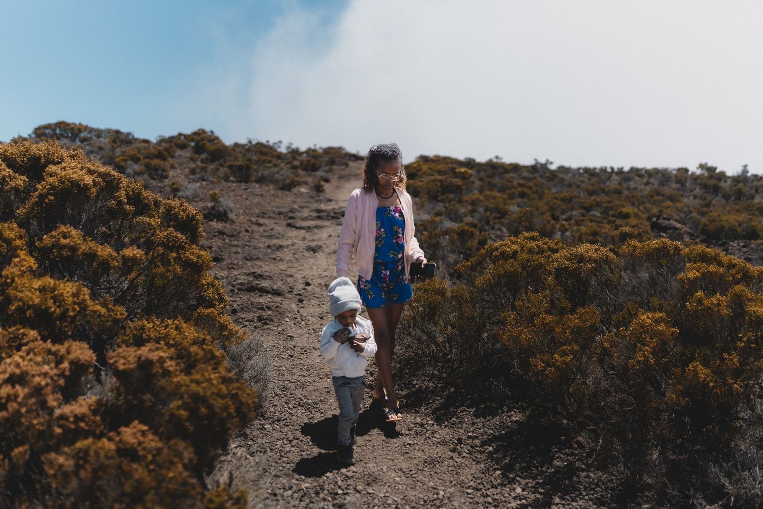 A woman and toddler hiking a rugged mountain trail surrounded by golden shrubs under a hazy sky.