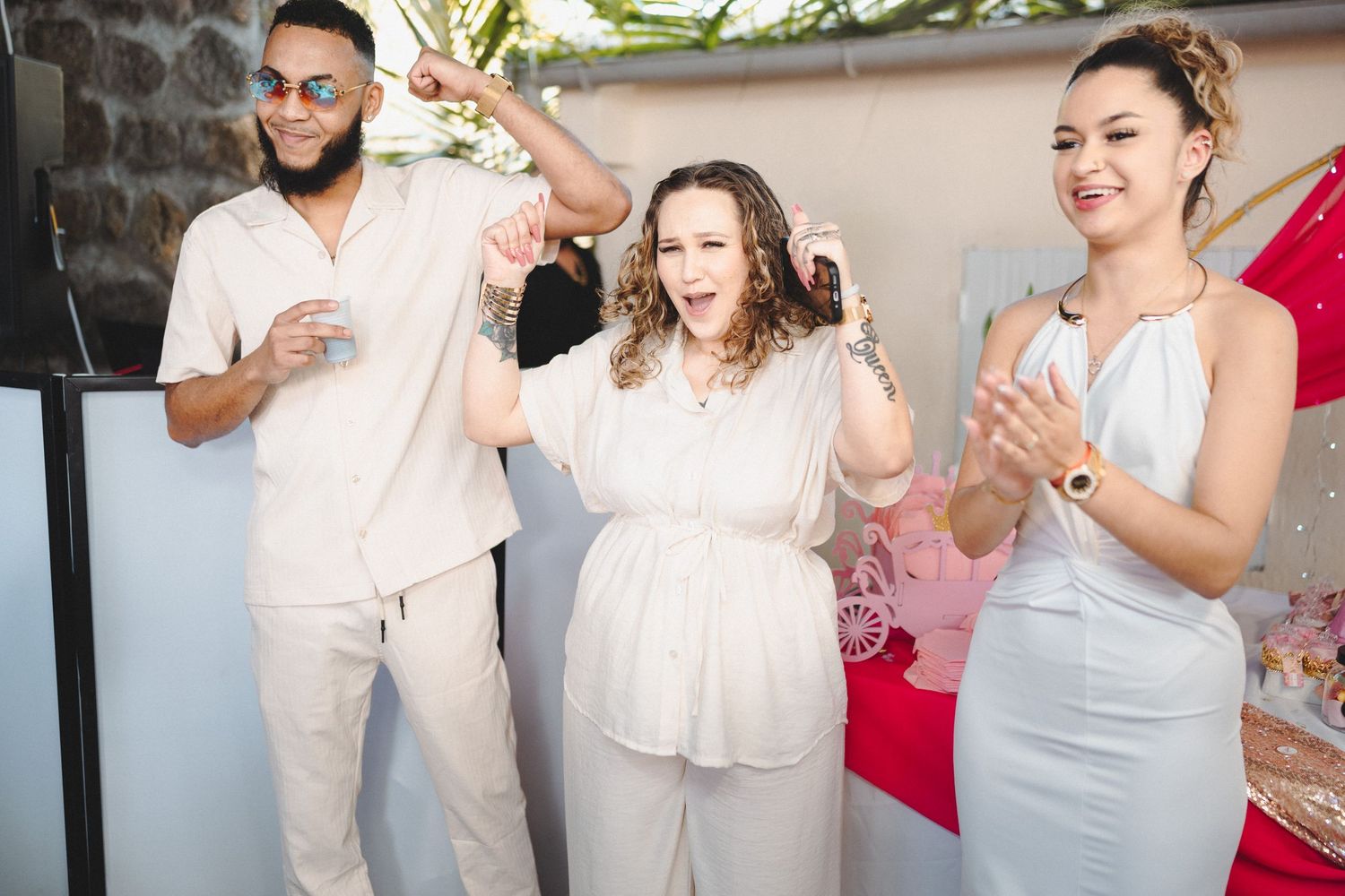 Three friends dancing and celebrating at an outdoor party, wearing white and light-colored outfits.