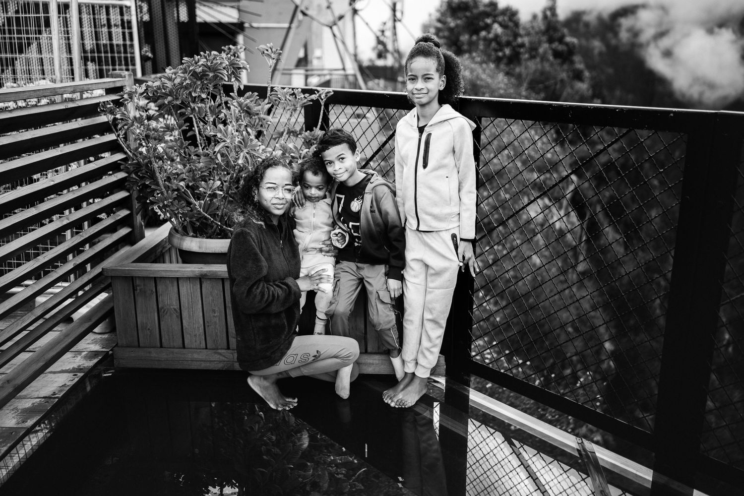 Black and white photo of a woman and two children posing together on an outdoor deck with plants and fencing.