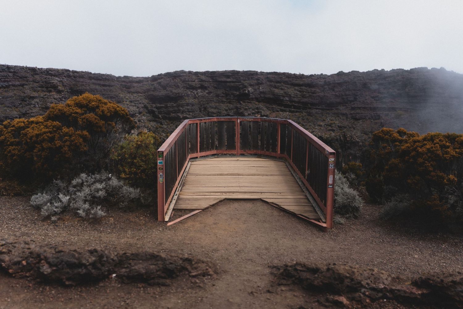 A rustic wooden bridge with red metal railings stands on a volcanic rocky landscape surrounded by sparse shrubs and fog.