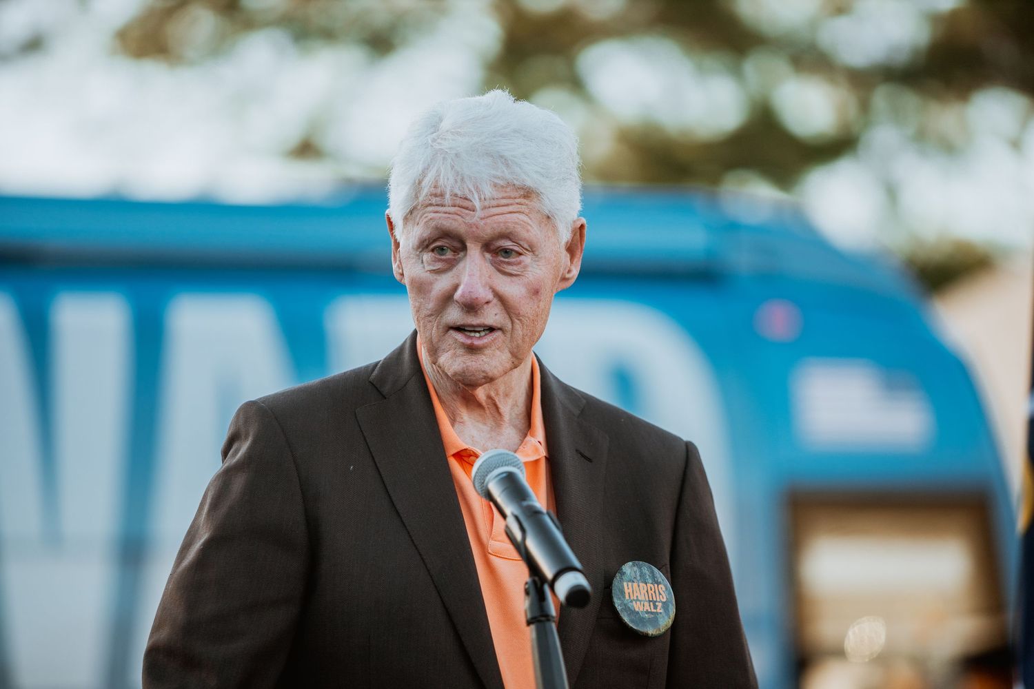 Elderly man with white hair in dark blazer and orange shirt speaks at outdoor event near microphone.