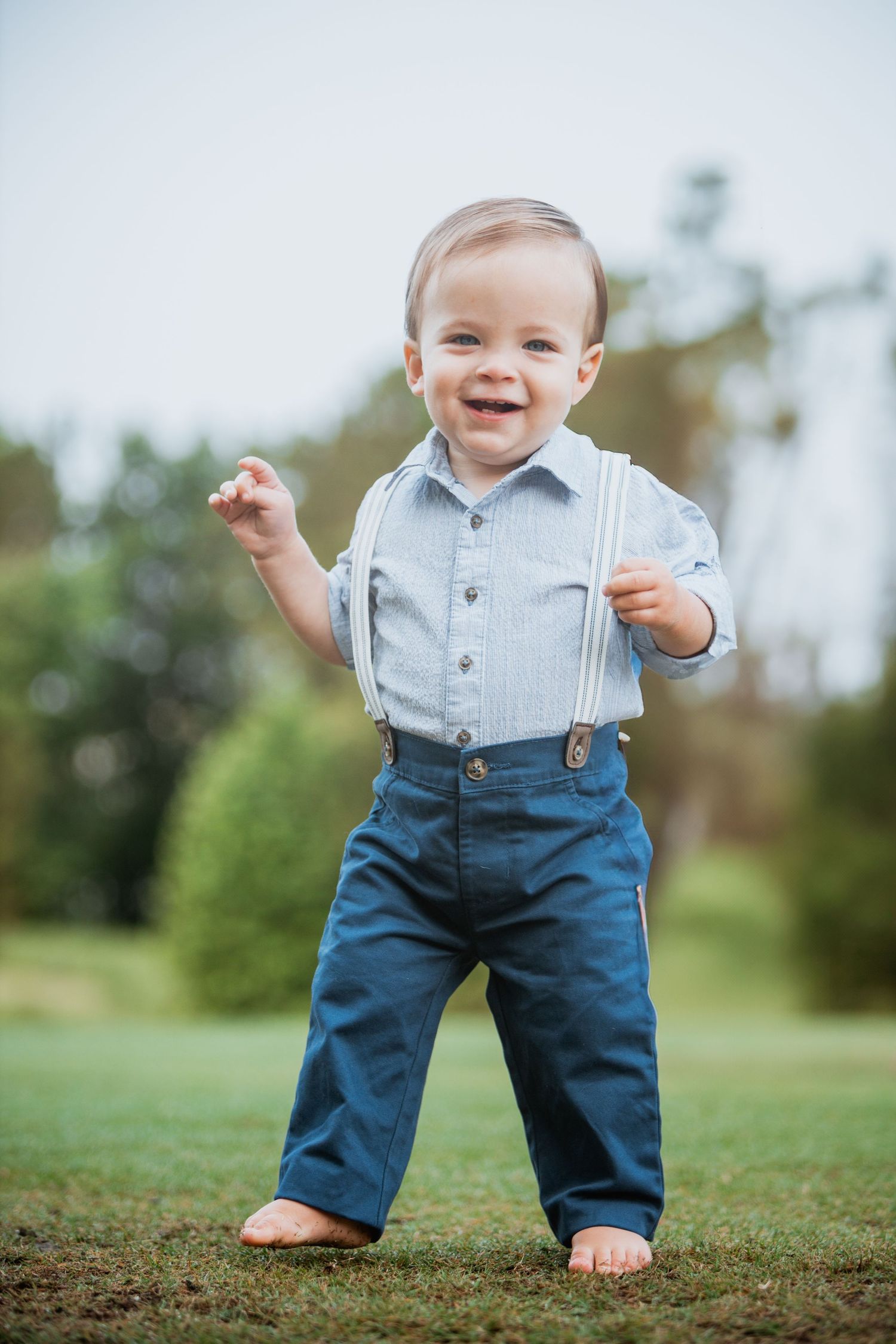 Happy toddler boy standing on grass wearing blue pants and suspenders in a sunny outdoor park.