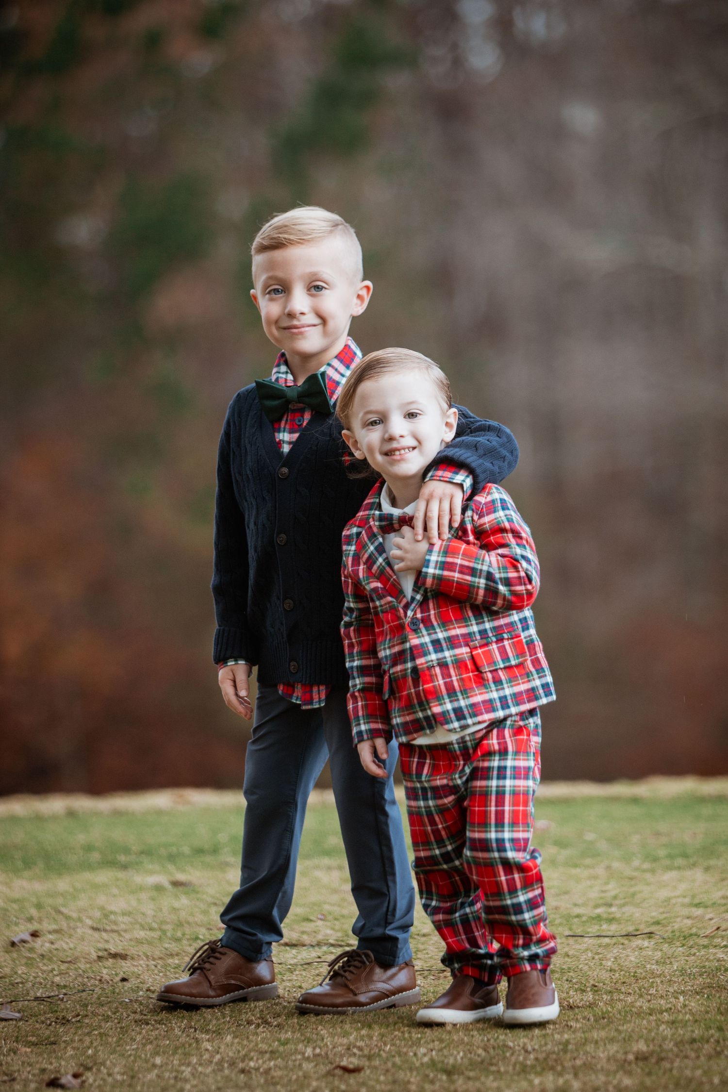 Two young brothers in coordinating holiday outfits pose together on a grassy lawn with fall foliage.
