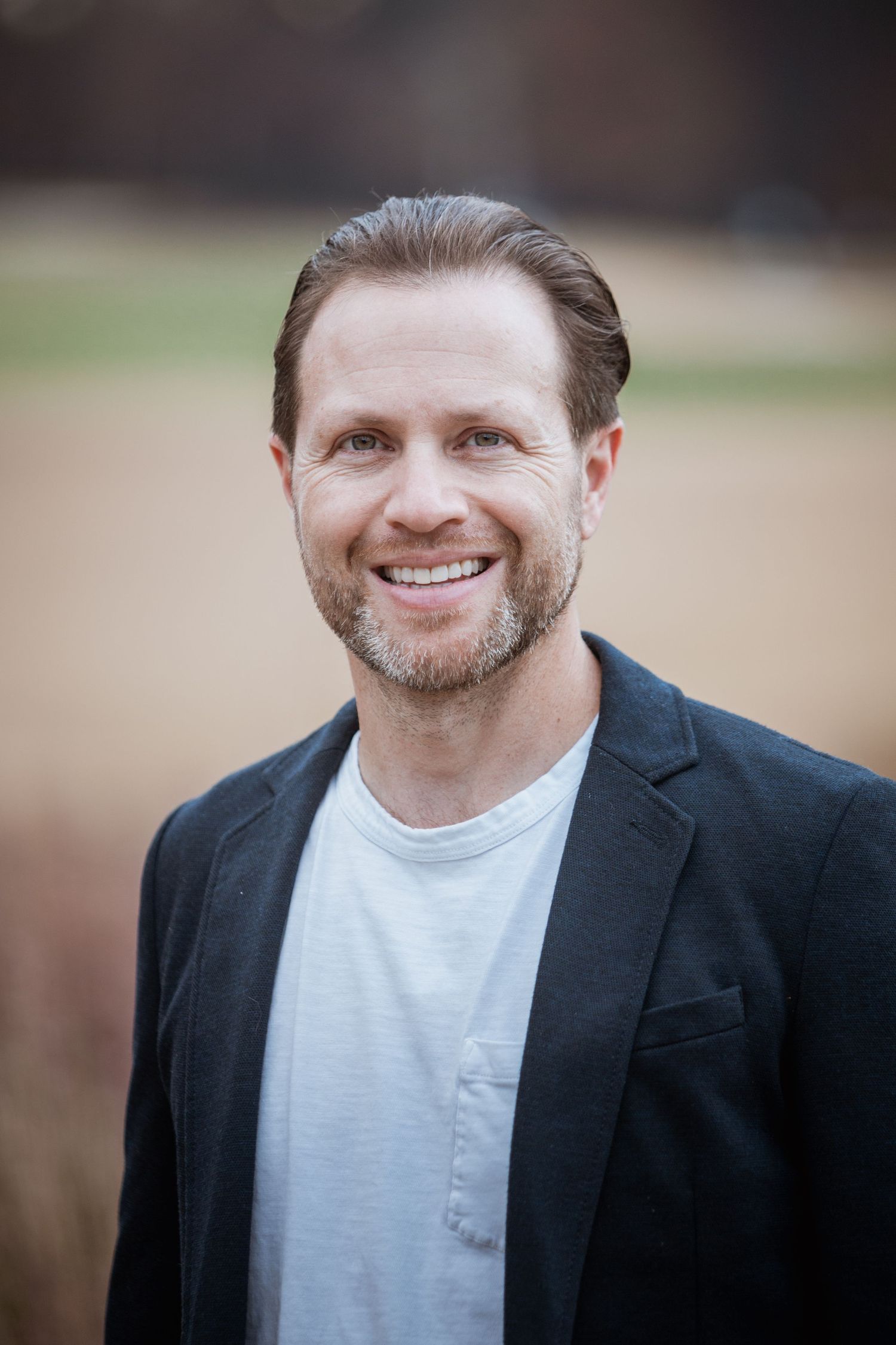 Smiling middle-aged man with beard in dark blazer and white t-shirt outdoors.