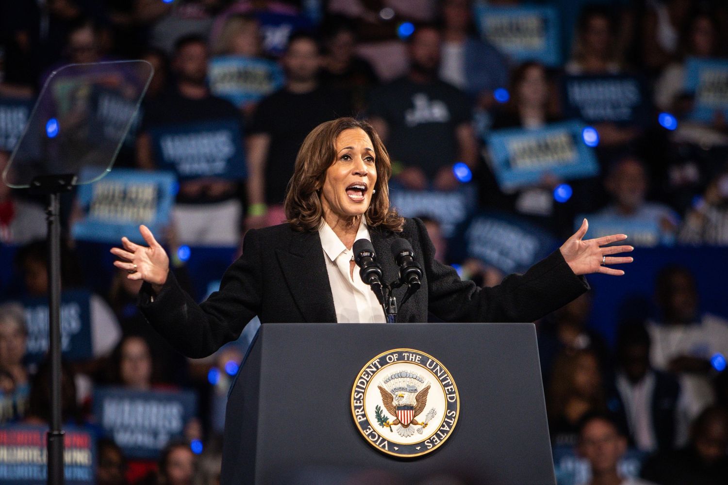 A woman in a black blazer spreads her arms wide while delivering a passionate speech at a campaign rally.