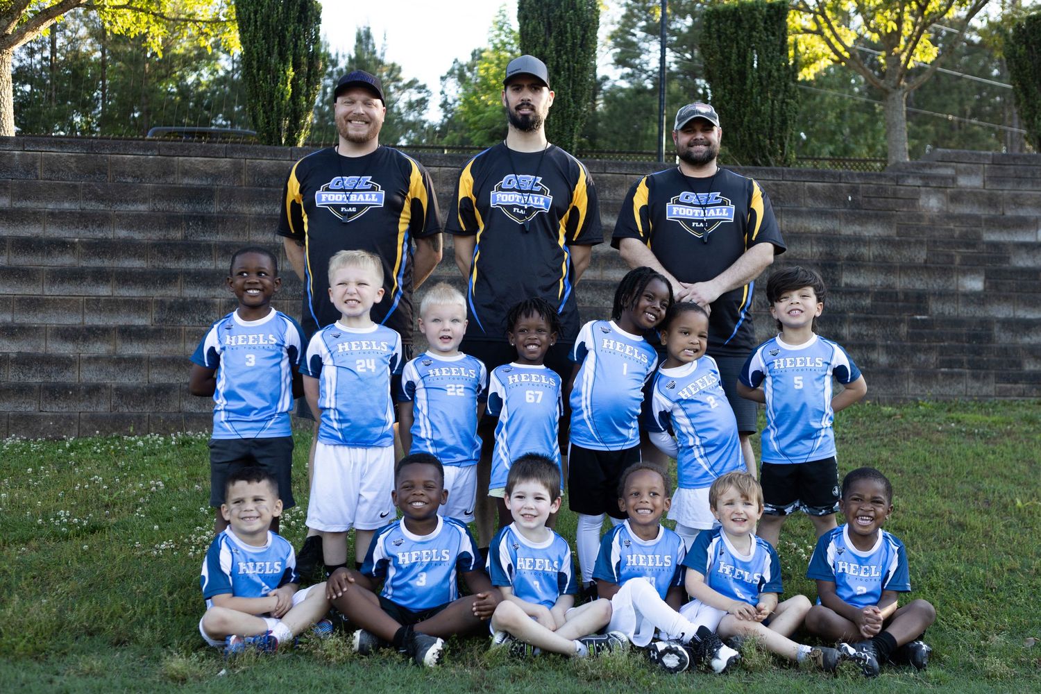 Youth flag football team in light blue jerseys poses with three coaches for outdoor team photo.