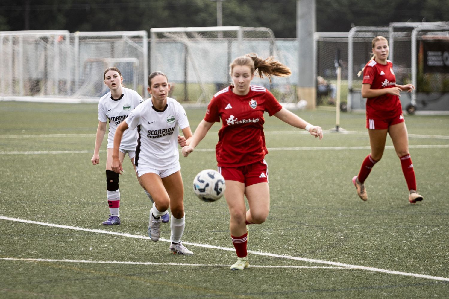 Female soccer player in red uniform chases ball ahead of white-kitted Scorpions defender during league match.