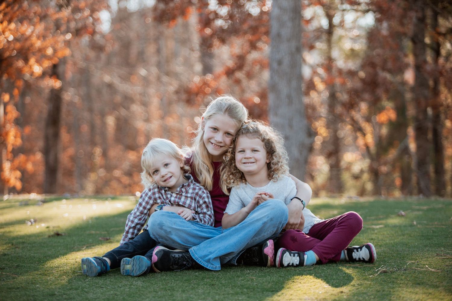 A smiling blonde mother sits on grass hugging her two young children with fall foliage backdrop.