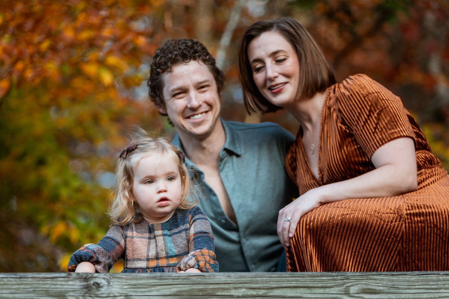 Happy family with curly-haired dad, brunette mom in rust velvet dress, and toddler daughter pose outdoors in fall foliage.
