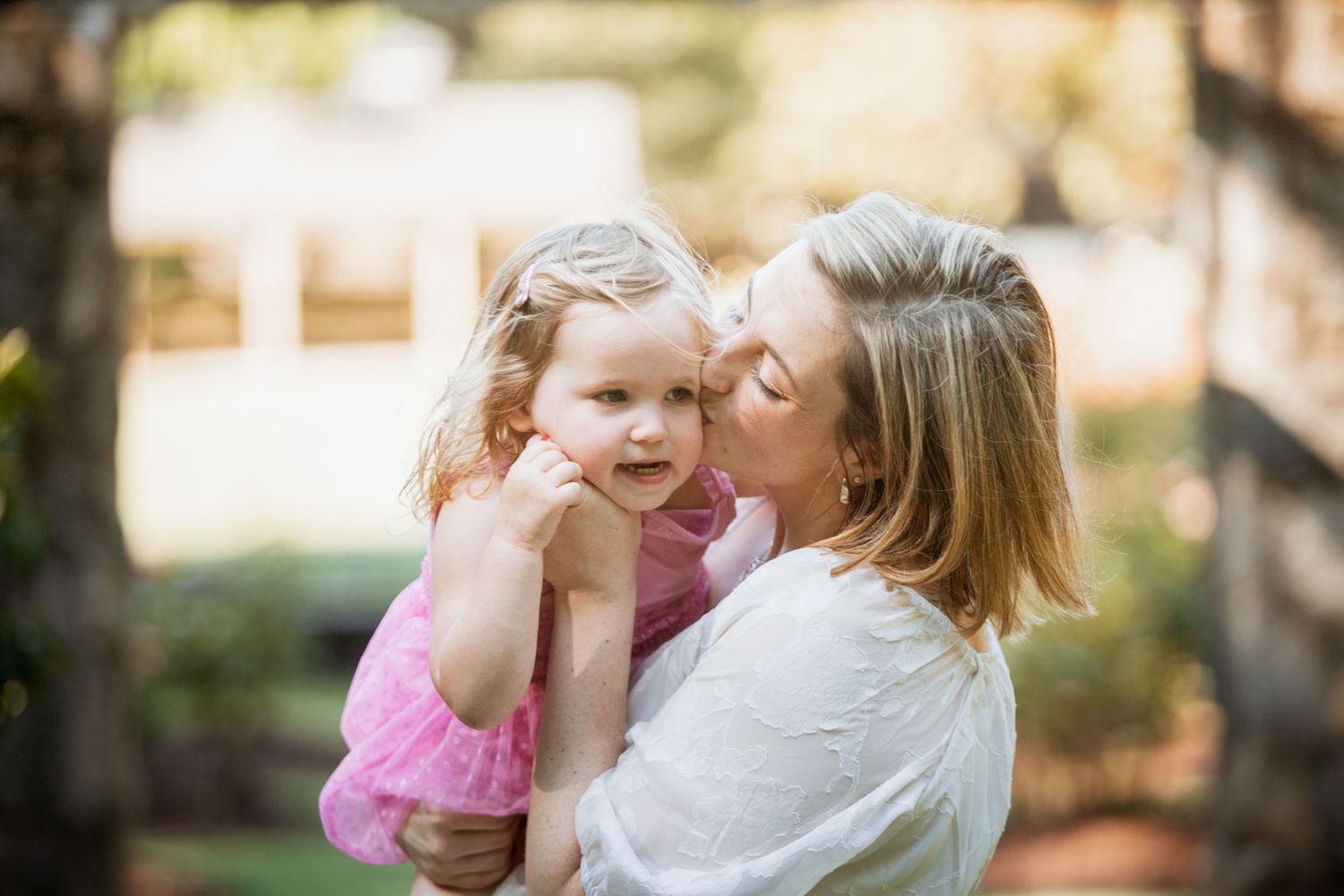 Mother tenderly kissing young blonde daughter on cheek in sun-dappled outdoor setting.