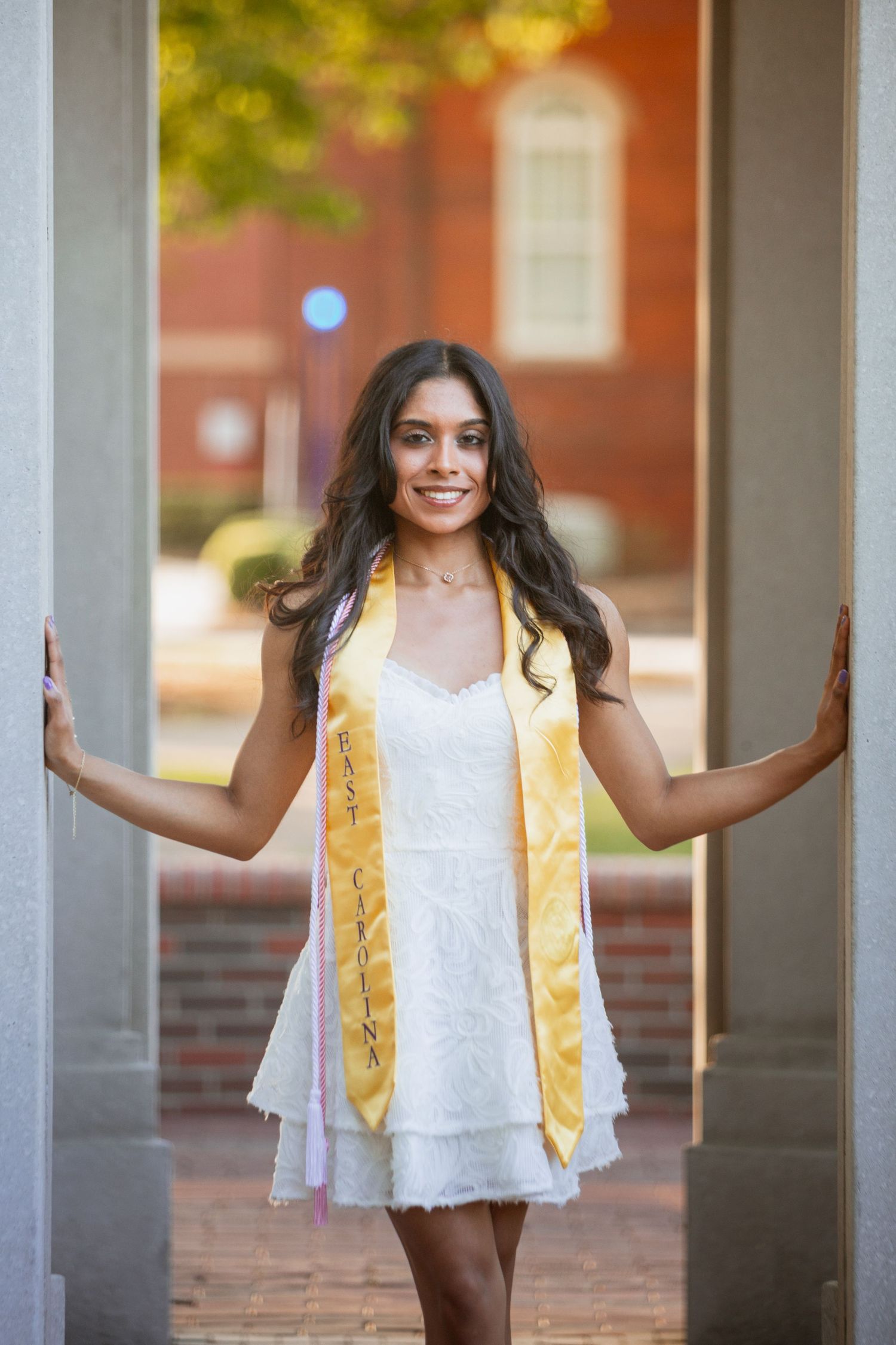 Graduate in white dress and East Carolina gold sash poses between two columns on campus.