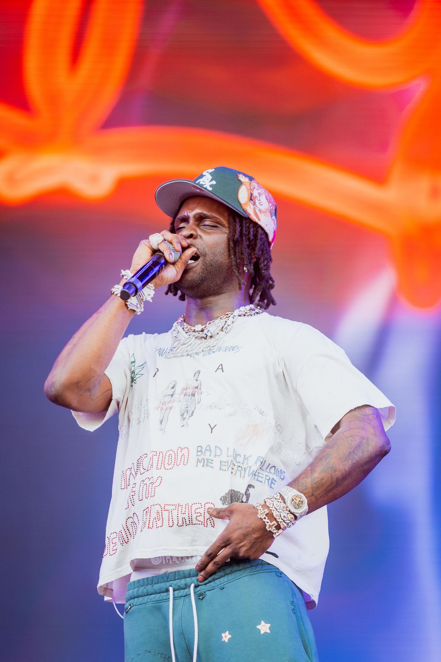 Rapper performing live on stage in a white graphic tee, teal pants, and layered chains under orange lights.