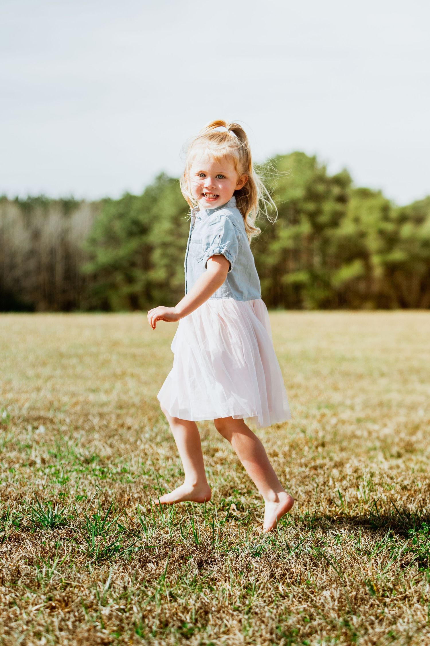 Happy blonde little girl in blue top and white tutu skirt running barefoot in sunny green field.