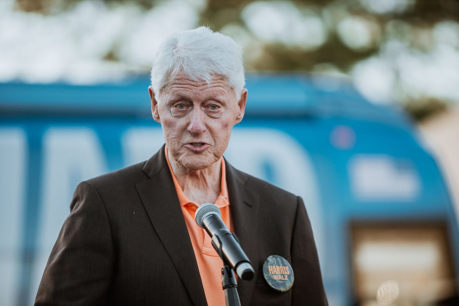 Elderly man with white hair speaking at a microphone outdoors, wearing a black blazer and orange shirt with a button pin.