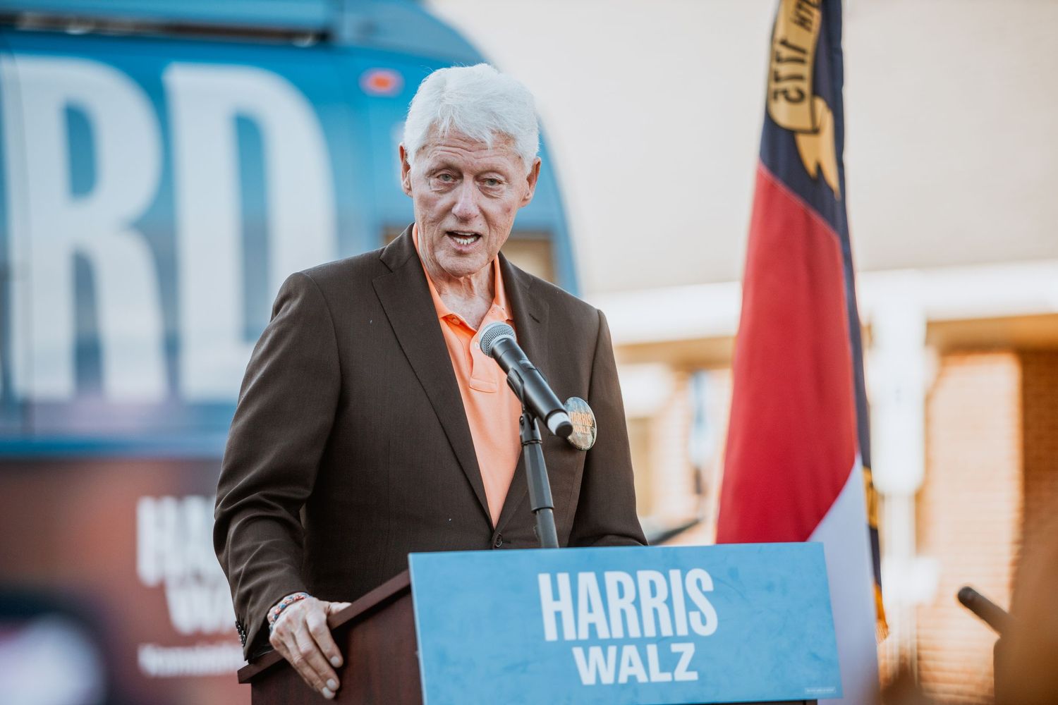 Speaker in brown blazer and orange shirt addresses crowd at Harris Walz political campaign rally.