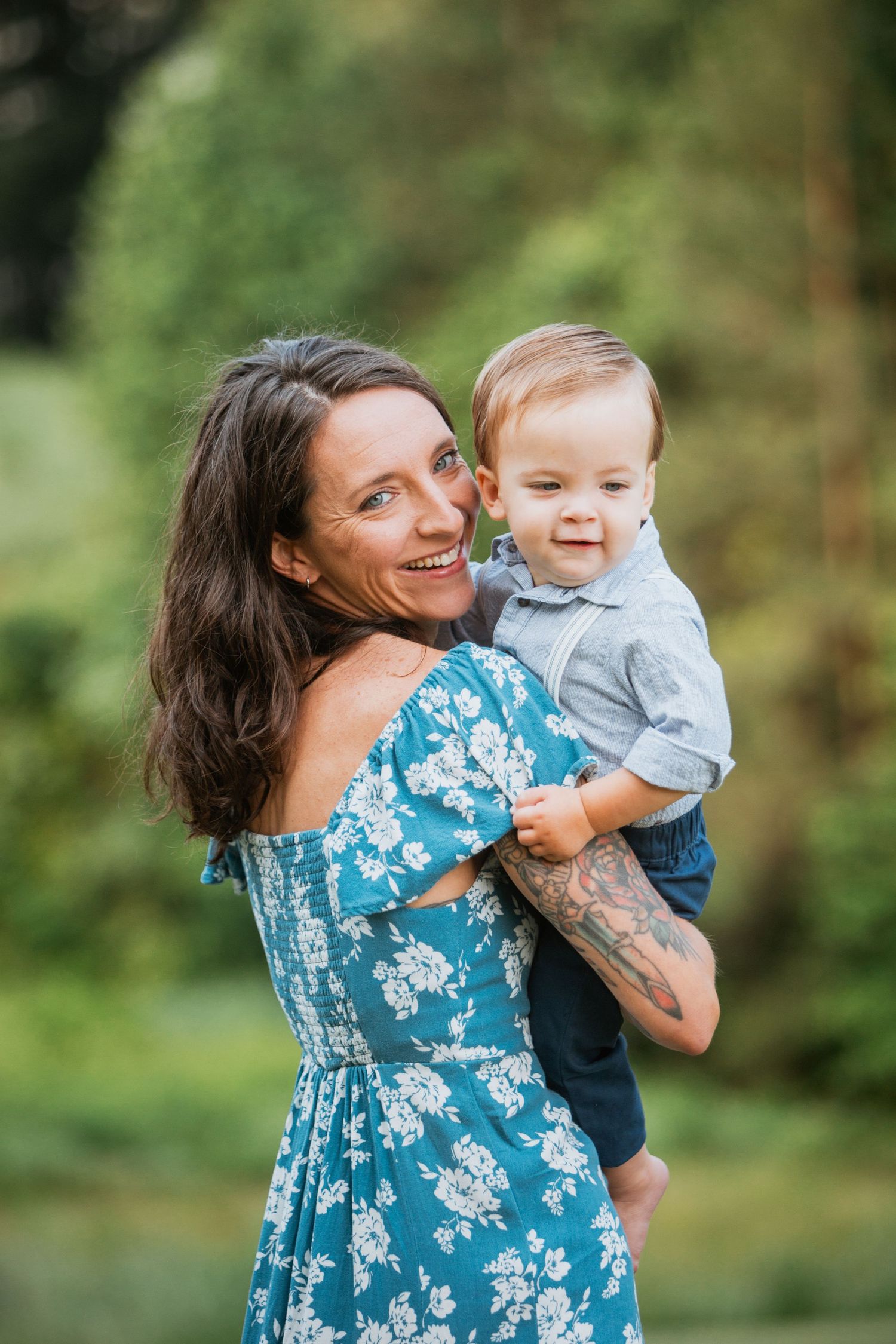 Smiling mother in floral blue dress holding her baby boy outdoors in a green natural setting.