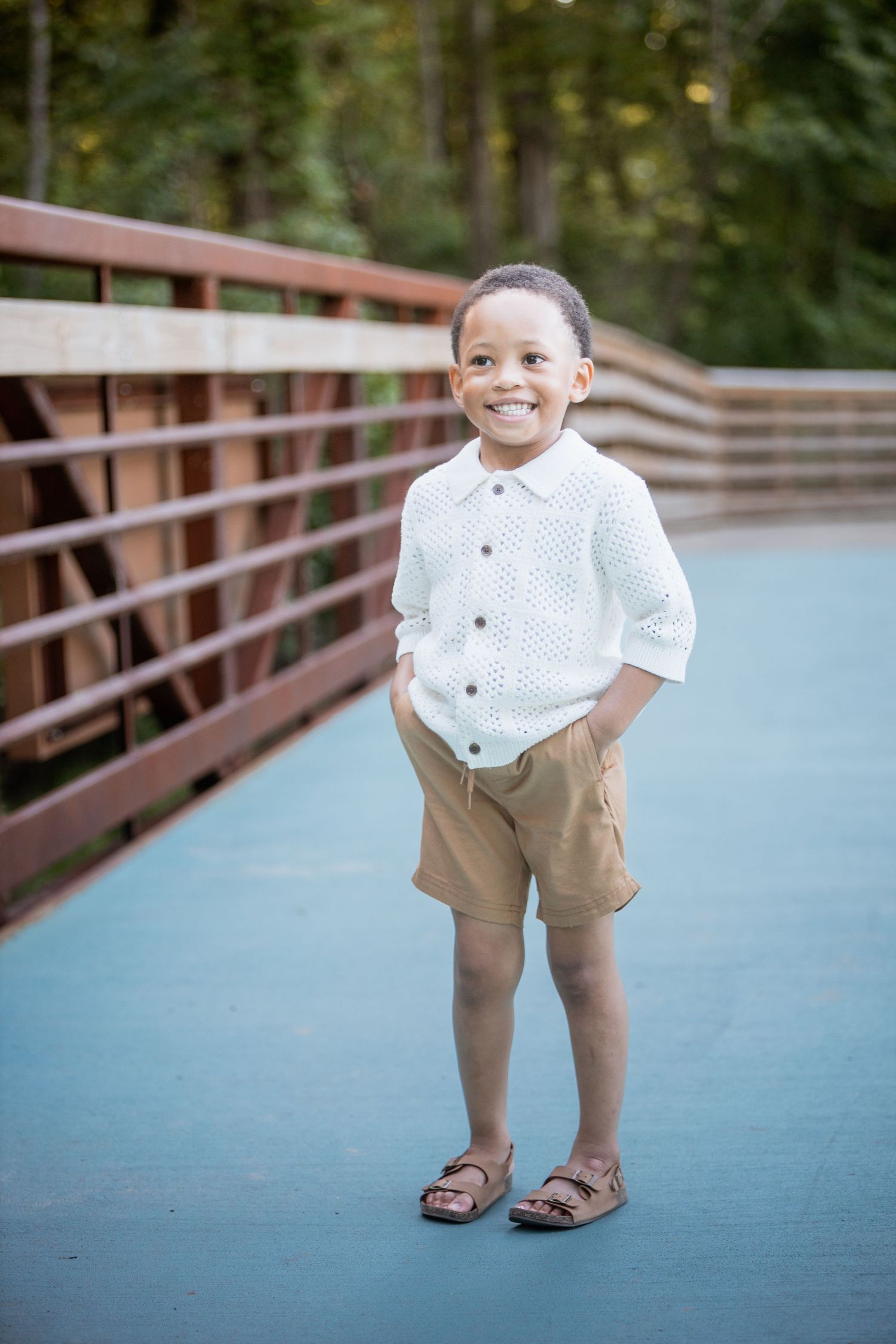 Smiling young boy in white knit shirt and khaki shorts standing on a blue bridge with hands in pockets.